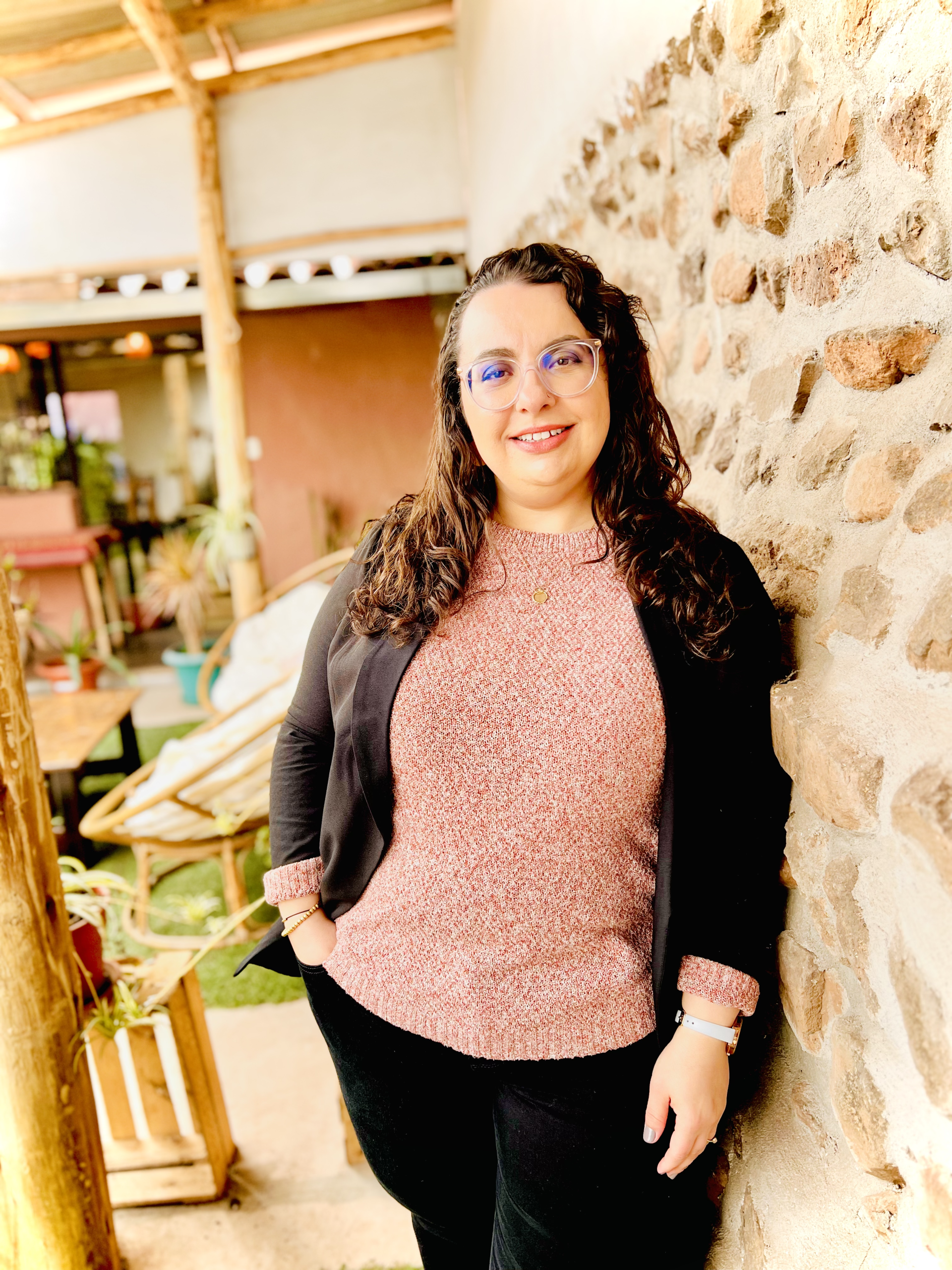 Nicole smiles confidently in a casual pink sweater and blazer, standing against a textured stone wall.