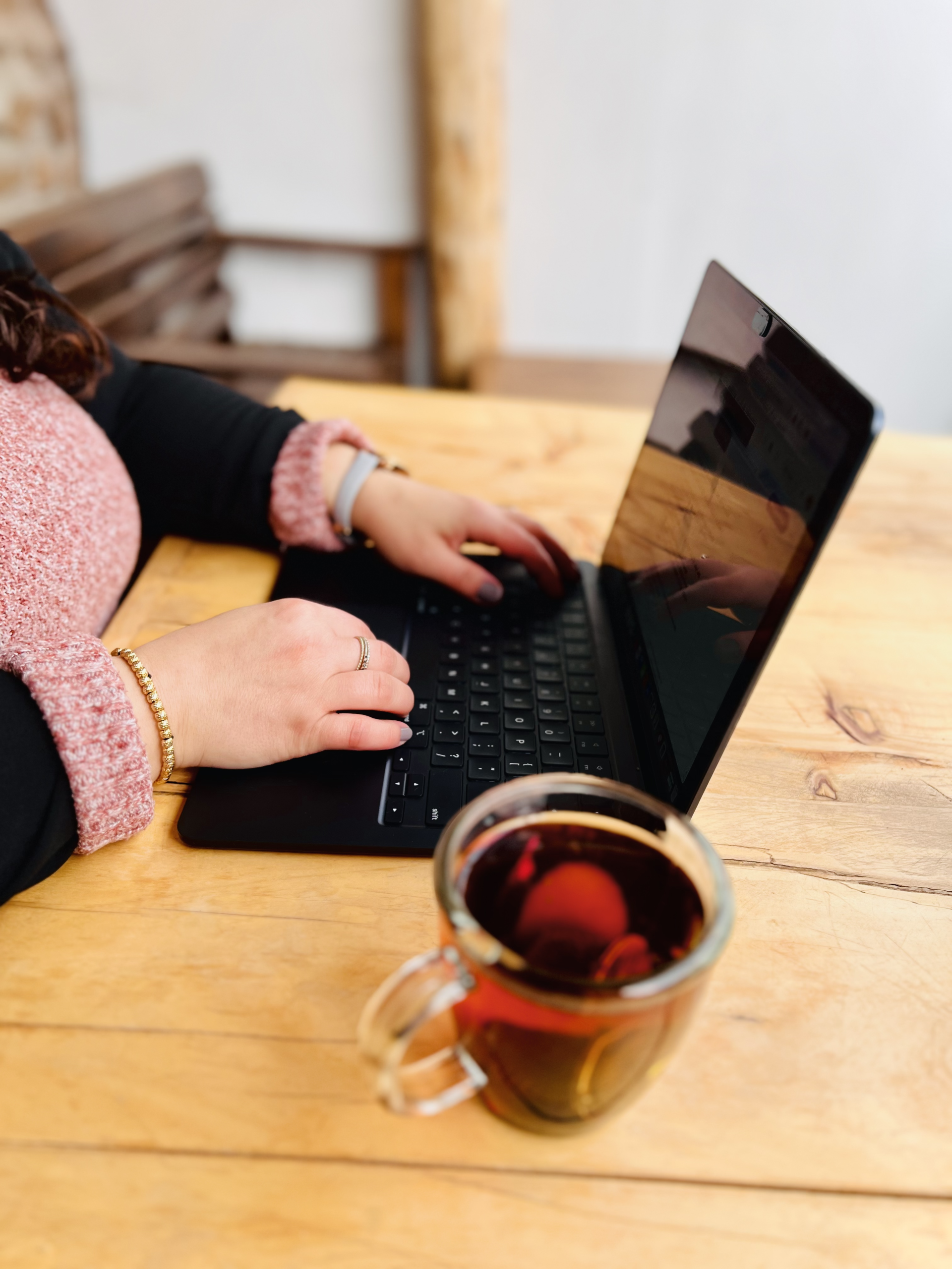 Close-up of a glass mug filled with black coffee on a wooden table, with a woman typing on a laptop in the blurred background. She is wearing a pink sweater and black blazer, creating a cozy and productive vibe.